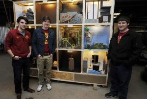 From left, Patrick Favo, Brendan Favo and Jeremy Price stand by the ISU Rube Goldberg Machine they made at Ames Golf and Country Club. The new Rube Goldberg Machine Club has spent the last semester building the 12 boxes of the machine, and will take the piece to a world competition later this month. (Photo by Nirmalendu Majumdar/Ames Tribune)