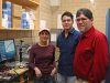 A faculty member, grad student and undergrad student stand together in a lab next to a instrument they built together.
