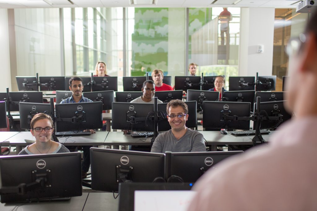An instructor faces a room full of students in a computer lab classroom