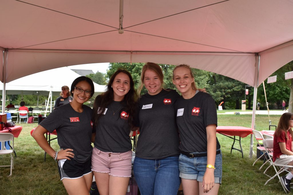 Four female college students stand in a white tent near picnic tables