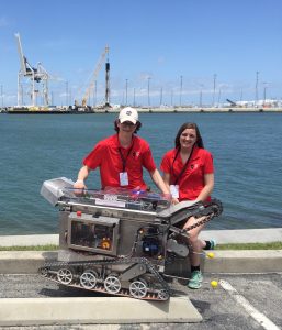 Two students stand in front of an ocean bay and pose behind a space mining robot they helped build