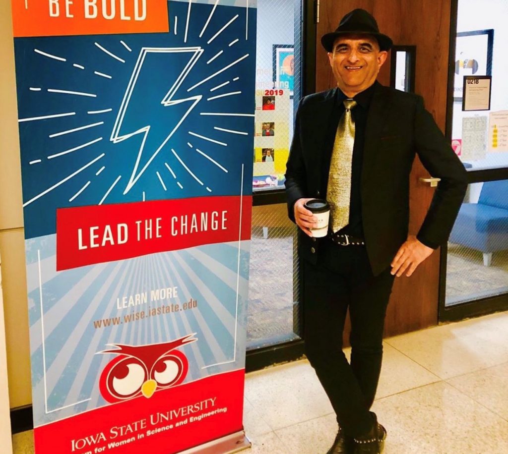 Man wearing a fedora hat and gold tie holds coffee in front of an office entrance