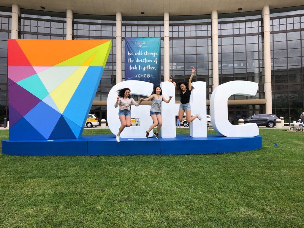 Three female college students are jumping mid-air in front of an outdoor sign with block letters