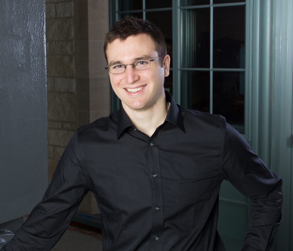 Ethan smiles while wearing a black collared shirt. A darkened window can be seen in the background.