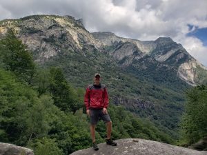 Former student stands on a large boulder with greenery and mountains in the background. Photo taken in Switzerland.