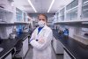 Scientist stands in a lab, wearing a white lab coat, gloves and a white face covering.
