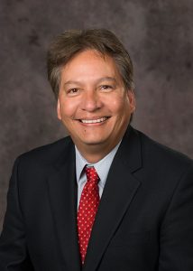 Headshot photo of Chris Cornelius smiling at the camera in a black suit with a red tie.