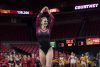 Courtney Middelkoop wears her gymnastics uniform which is black on the bottom and maroon on the top. Courtney is smiling with her hands over her head. Her teams and cheerleaders cheer her on in the background inside Hilton Coliseum.
