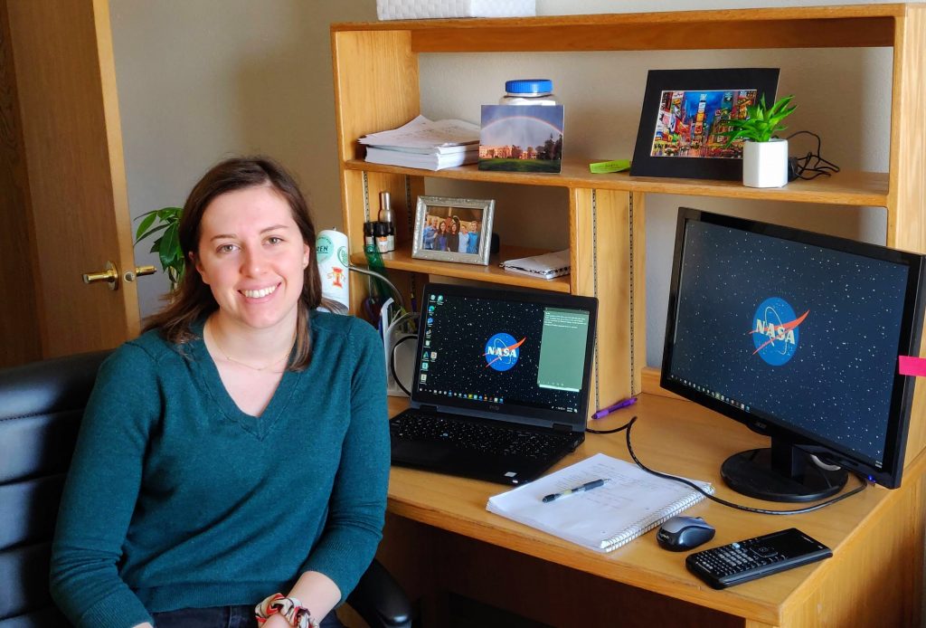 Mechanical engineering student Olivia Tyrrell smiles and poses in front of her desk. Both of her computer monitors have the NASA logo on them. A notepad and calculator are also seen in the shot.