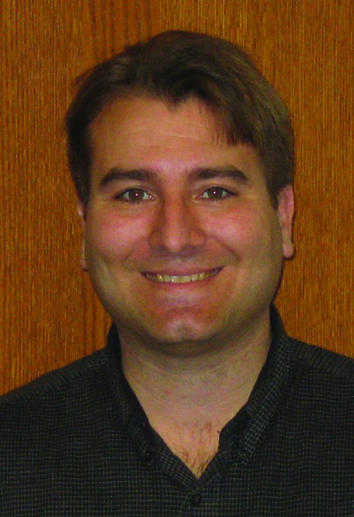 Mechanical engineering professor Michael Olsen smiles as he poses in front of a wooden door. He is wearing a black collared shirt. 