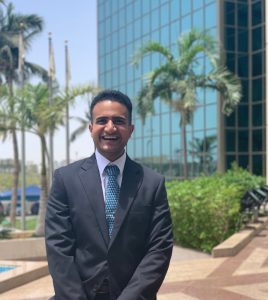 A man wearing a suit and tie smiles. In the background is a large building with lots of windows and various palm trees.