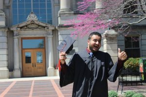 A man in a graduation gown smiles and gives a thumbs up. He's holding his graduation cap. In the background is Marston Hall, a bedroom limestone building on the Iowa State University campus.