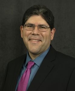 Mechanical engineering assistant professor Jaime Juarez smiles while wearing a dark suit jacket, a blue collared shirt and a red neck tie