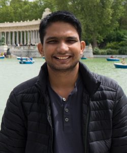 Mechanical engineering graduate student Ankush Mishra poses while wearing a black jacket and a blue collared shirt. Boats on water can be seen in the background.