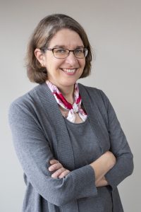 Sarah Ryan smiles and crosses her arms. She's wearing a grey sweater, a grey blouse, and a colorful scarf.