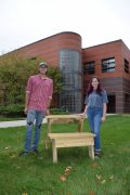 Two students stand outside of Black Engineering Building next to a desk they helped to design and fabricate.