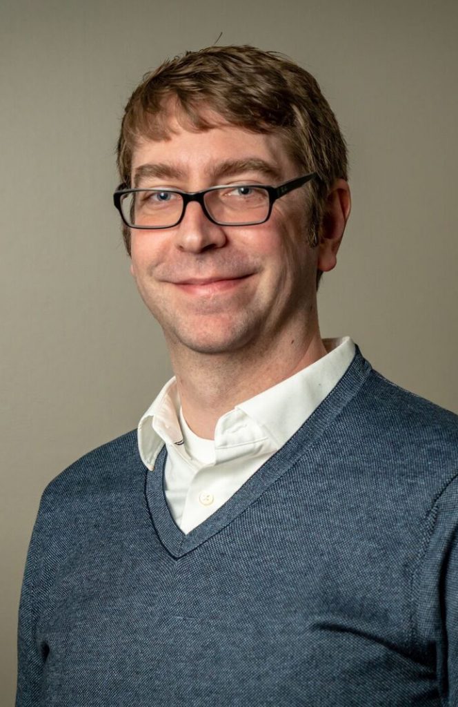 A headshot of Christopher Whitmer. He is wearing a blue v-neck sweater and a white collared shirt.