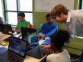 Chris Whitmer stands over a student's shoulder and helps them use his software on laptop. Two other students are in the background of the shot on their own computers.