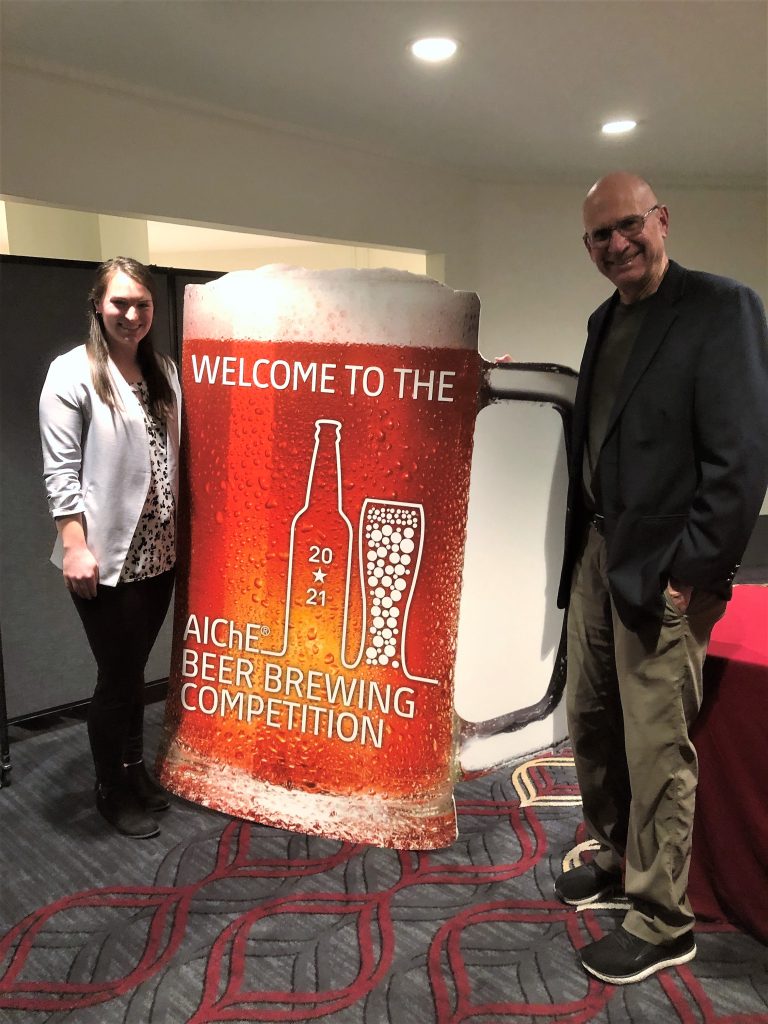 A female student and a male professor stand next to a giant cutout of a beer mug with the text: "WELCOME TO THE 2021 AIChE BEER BREWING COMPETITION