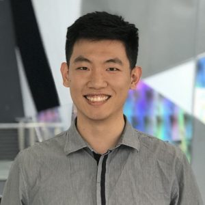 A student smiles for the camera. He is wearing a grey collared shirt