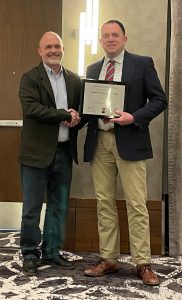 Two men stand together shaking hands. One of the men is holding the certificate for an award he won.