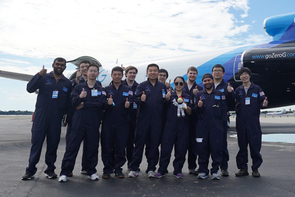A group of Iowa State University engineering researcher pose in front of an aircraft and give thumbs-up to the camera. All researcher are wearing blue jumpsuits.