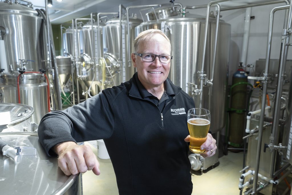 A man poses with a beer. Brewing equipment can be seen in the background.