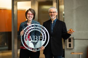 Photo of Department Chair Amy Kaleita and professor Jacek Koziel holding two retirement gifts for Koziel: A carved metal sign with his name and a glass globe with cyclone colors inside