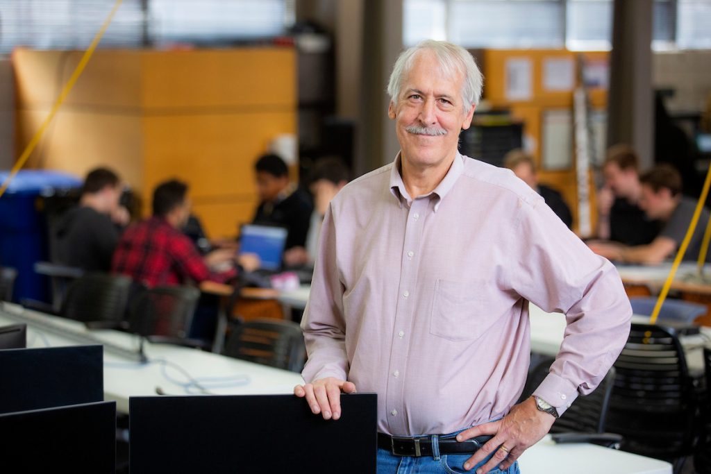 Professor Doug Jacobson standing and smiling in a classroom with hands on his hips. 