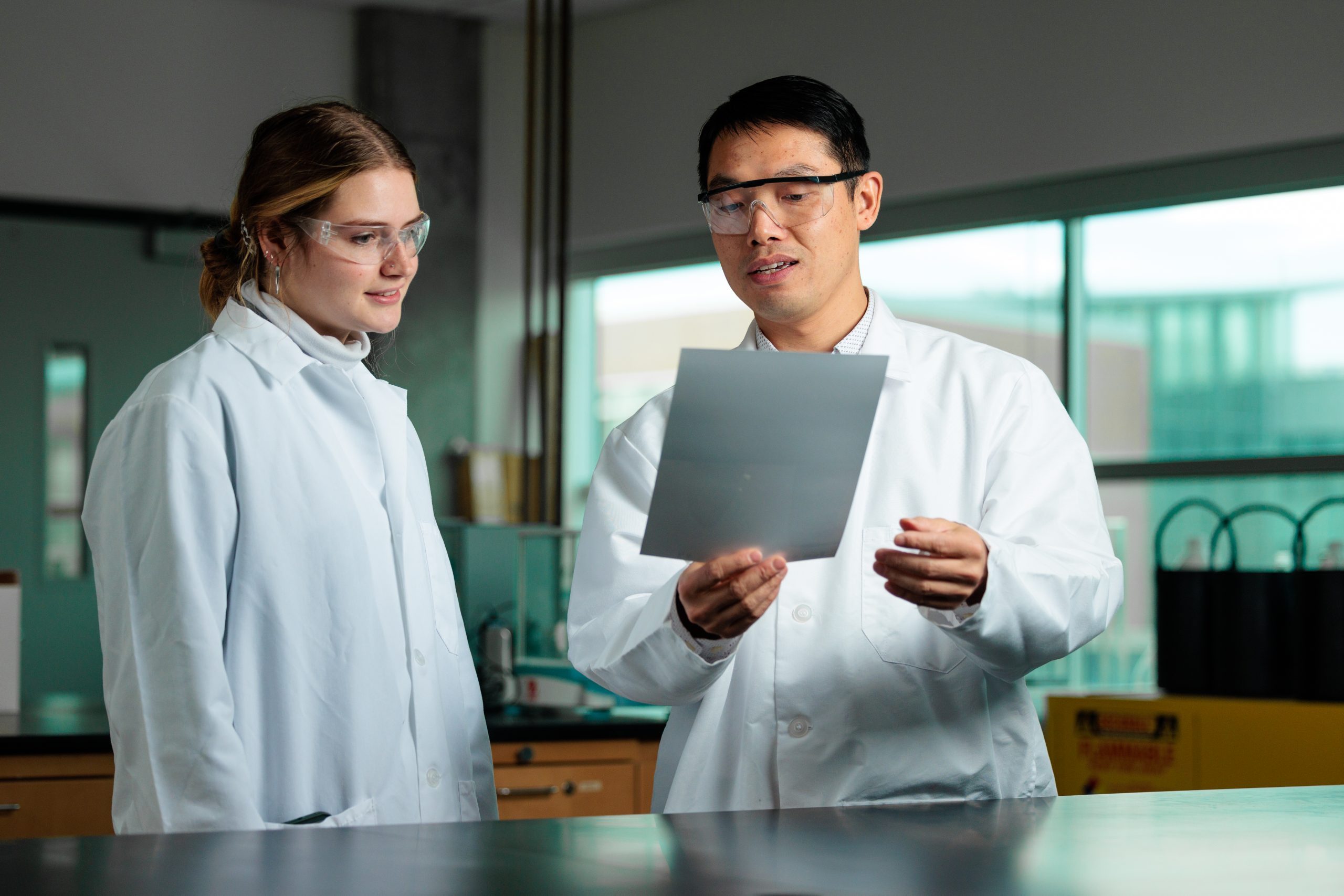 Dr. Shan Jiang standing and speaking to a student next to him. Both are looking at a document. 