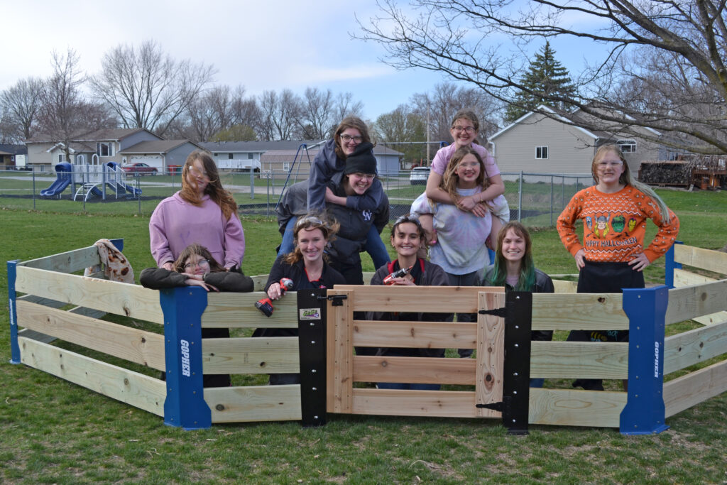 Abby McCormick, mechanical engineering major, and a group of girls smile at the camera from inside a gaga ball pit enclosure they are assembling with drills.
