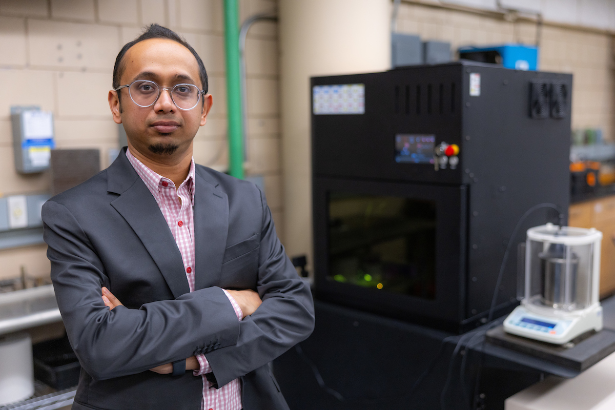 Roy Sougata stands with arms crossed in front of 3D printer in lab.