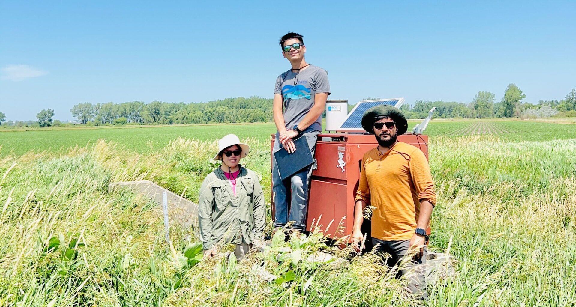 Three researchers smile in a field of grass, collecting water quality data.