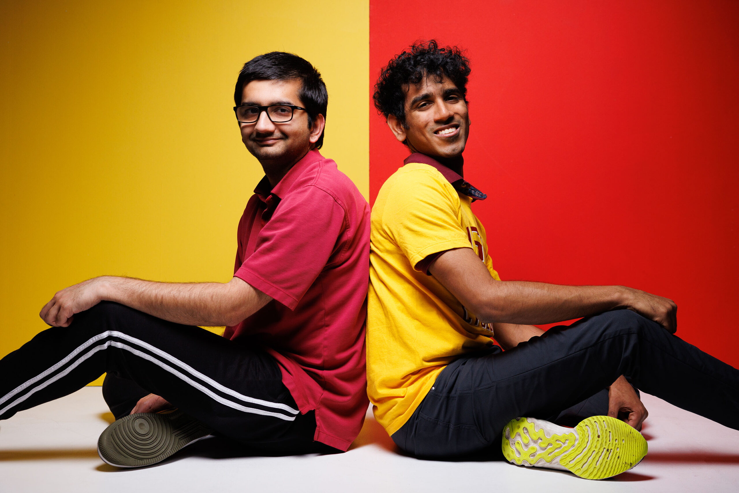 Deep Patel, left, and Dhananjay Dileep sit back-to-back and pose for a portrait in front of a cardinal and gold backdrop.