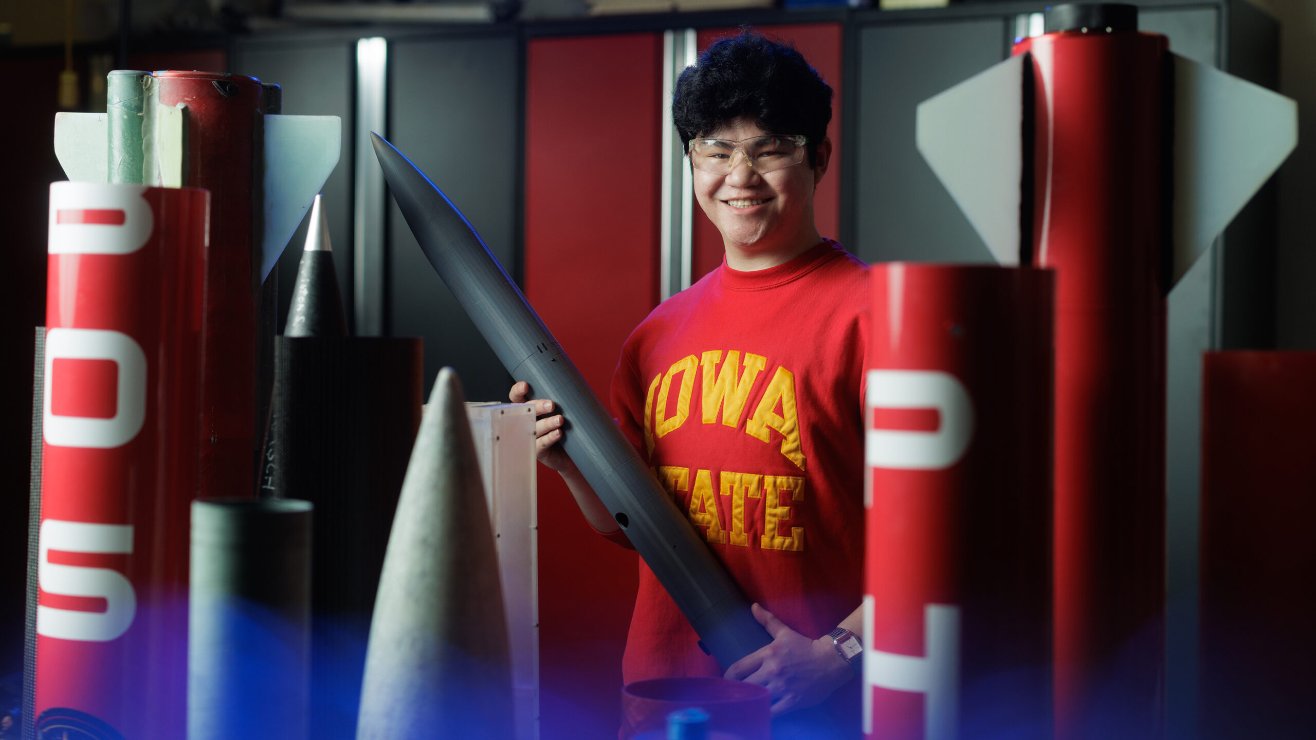 Gabriel Hoehl poses for a portrait in an Iowa State sweatshirt, surrounded by rocketry equipment with a blue light in the foreground.