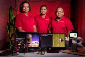 Three professors wearing red shirts looking at the camera smiling in front of computer screens of their AI digital twins.