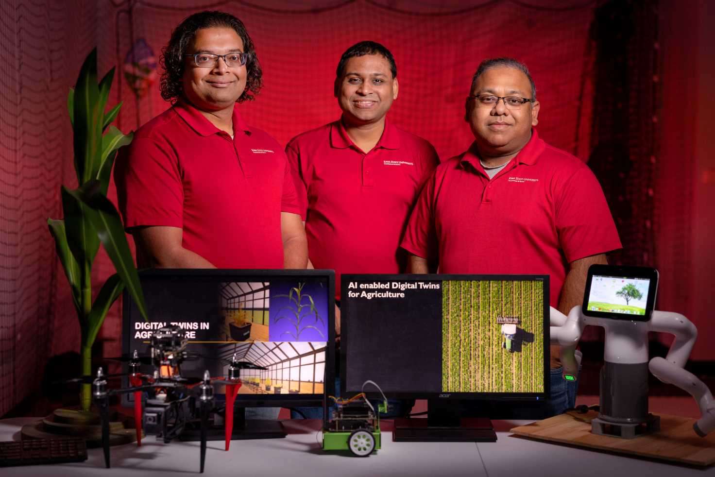 Three professors wearing red shirts looking at the camera smiling in front of computer screens of their AI digital twins.