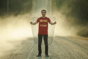 Araz Hasheminezhad holds a large piece of geogrid, arms-length wide and from his knees to his chin, while standing on a gravel road with dust accumulating around him.